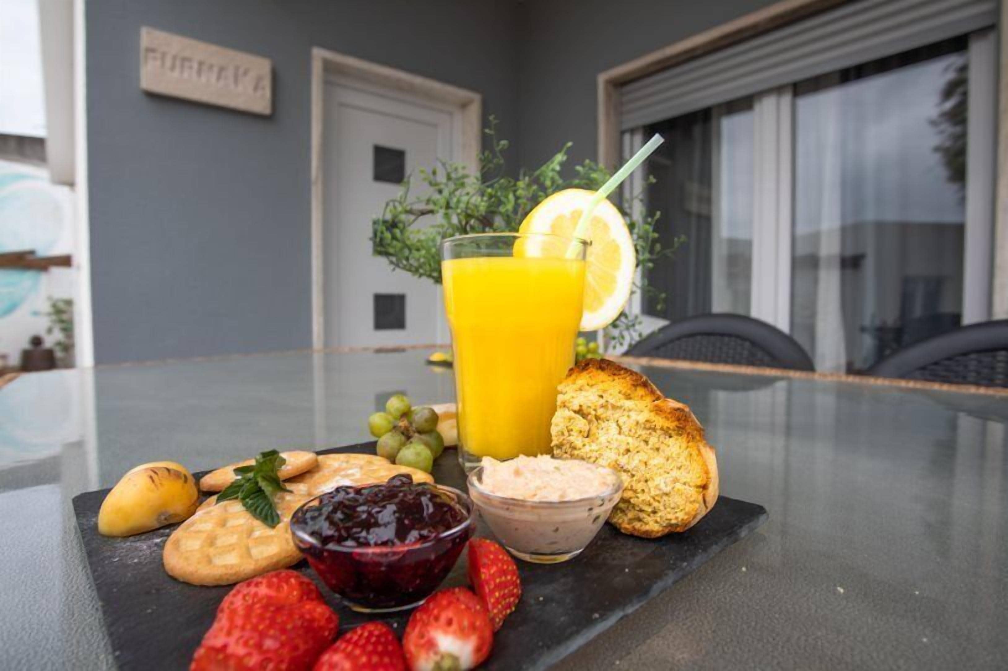 Una mesa con desayuno: tostadas, fruta, mermelada y un vaso de jugo de naranja.