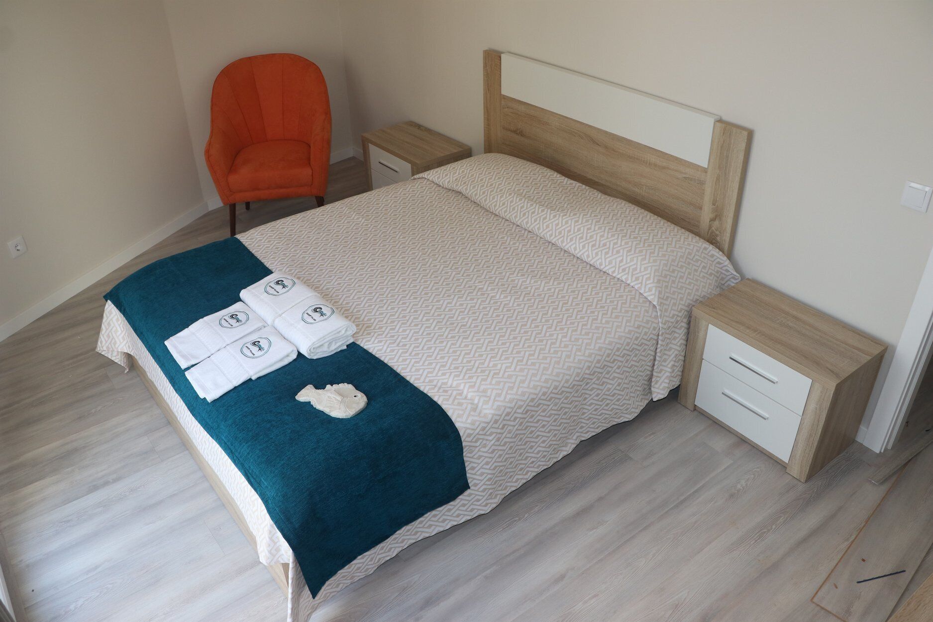 A neatly made bed with a patterned bedspread and a teal blanket, two bedside tables with drawers, and an orange chair.