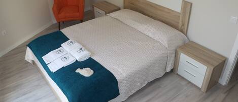A neatly made bed with a patterned bedspread and a teal blanket, two bedside tables with drawers, and an orange chair.