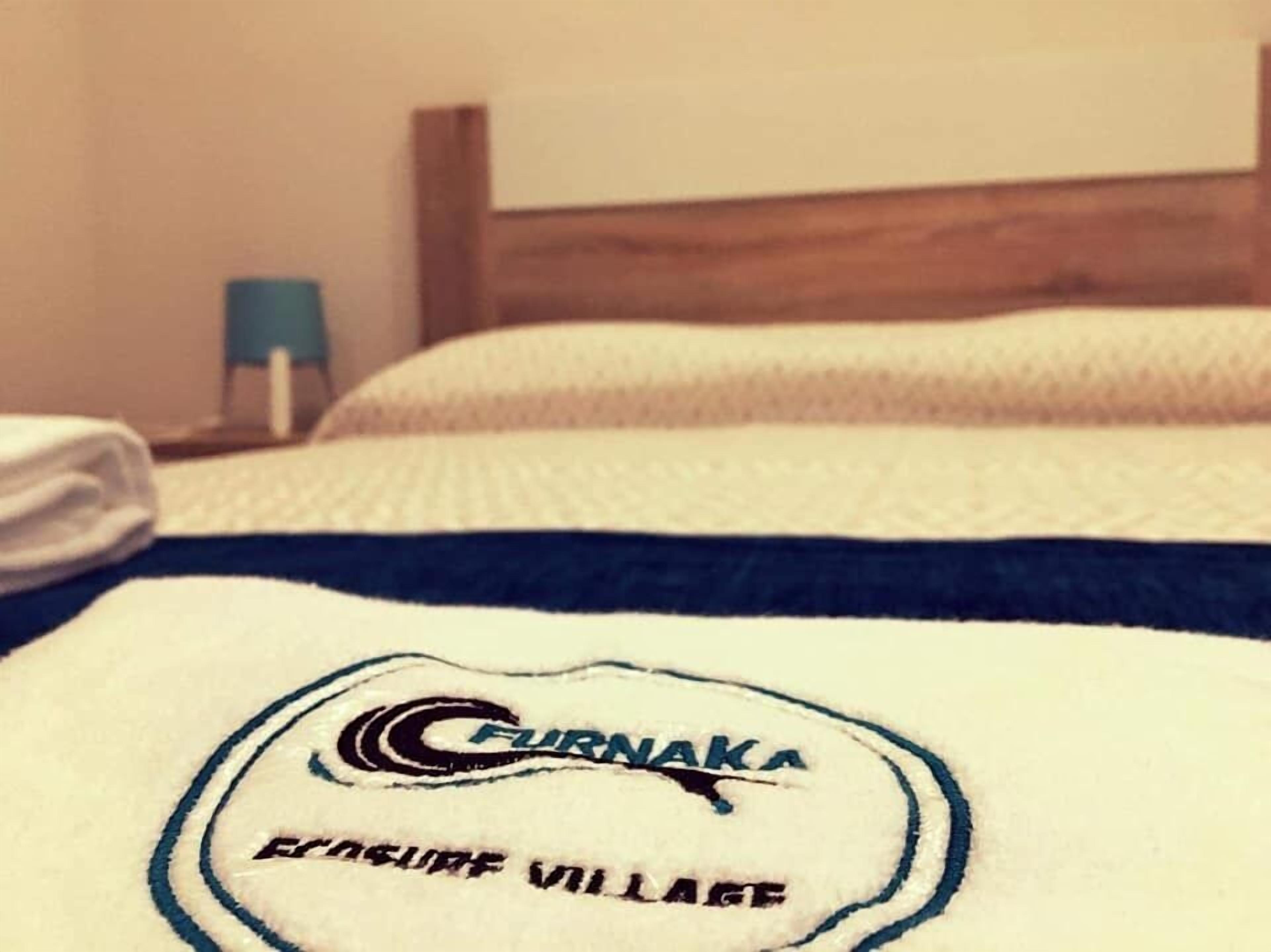 A bed with a white and blue blanket featuring the FURNAKA logo and the text "EASTSIDE VILLAGE".