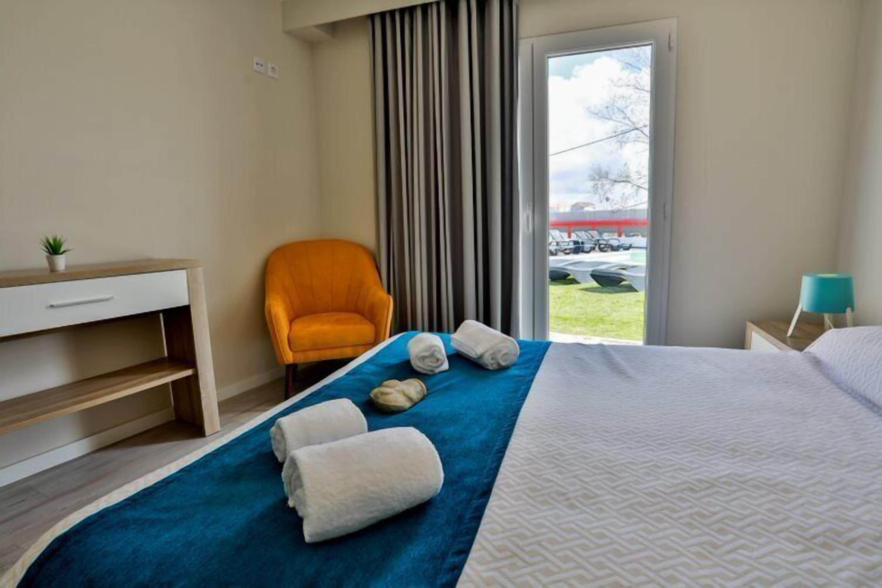 A hotel room with a bed, a desk, an orange chair, a window with curtains, and a view of the outside.