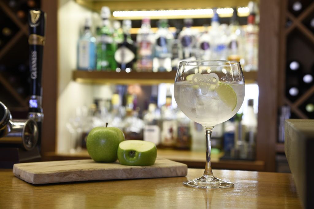 A bar setting with a glass of a clear beverage, a whole and a half-cut apple on a wooden board, and a bar counter with various bottles in the background.