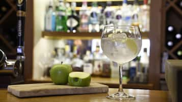 Un bar con una copa de bebida clara, una manzana entera y media cortada sobre una tabla de madera, y una barra con varias botellas al fondo.