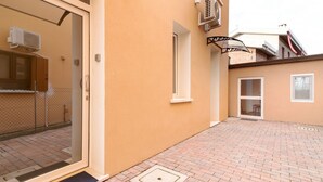 Un edificio con entrada por puerta corredera de vidrio, una ventana pequeña y un patio empedrado con ladrillos.