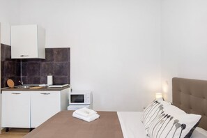 A compact hotel room with a kitchenette, a bed with a brown headboard, and a bedside lamp.