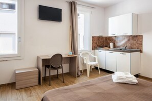 Een slaapkamer met een bed, een bureau, een stoel, een televisie, een kitchenette en een raam.