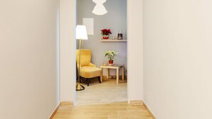 Ein Flur mit einem gelben Sessel, einem kleinen Tisch mit einem Topfpflanzen und einem an der Wand befestigten Regal mit Dekorationen, sowie einer weißen, engelhaften Silhouette.