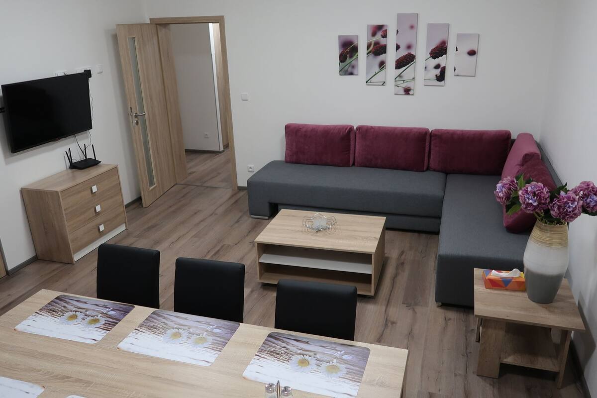 A modern living room with a grey sofa, a wooden dining table with chairs, a TV mounted on the wall, and a small side table with a vase of flowers.