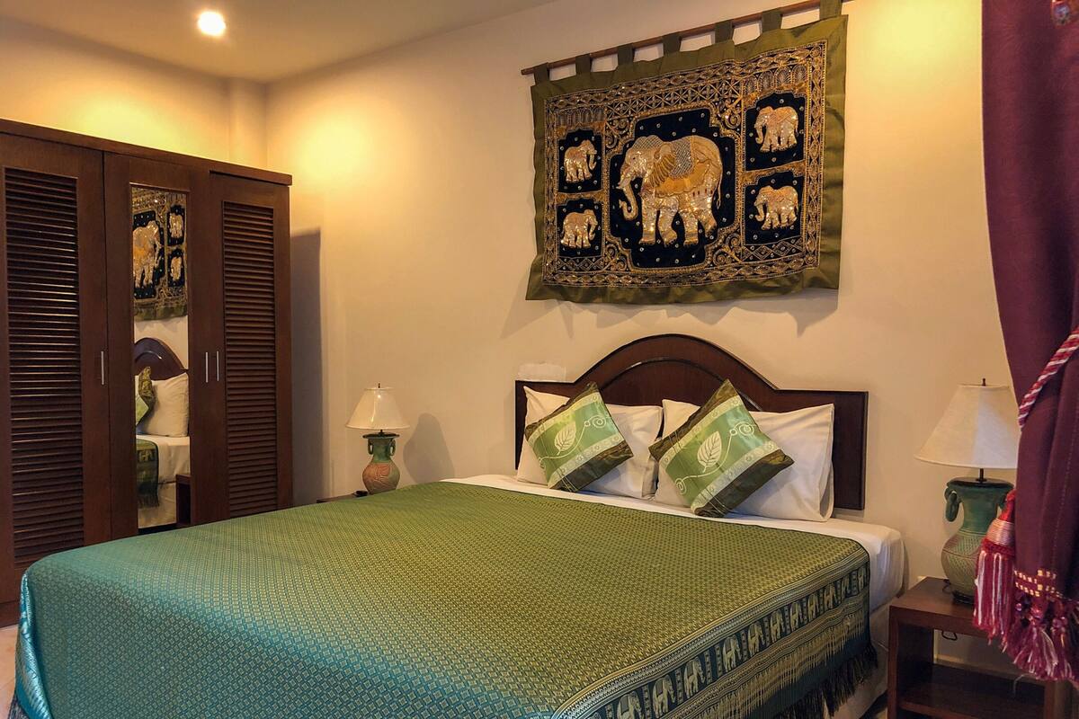 A neatly made bed with a green bedspread, two pillows, a headboard, and a wall tapestry with elephant designs.