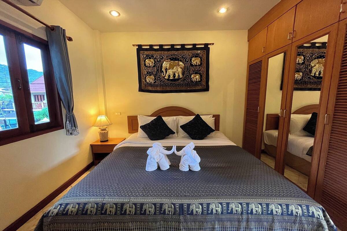 A hotel room with a bed, a nightstand, a lamp, a window with curtains, a mirror, and a wall tapestry.