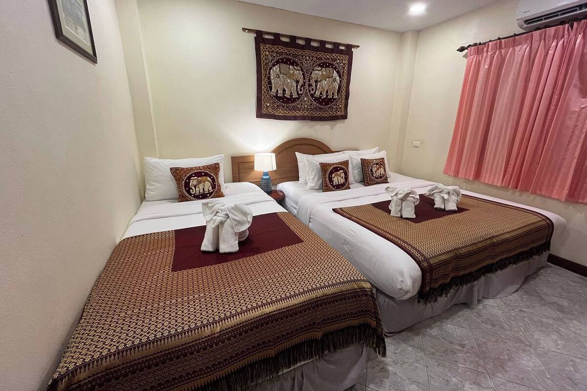 A hotel room with two beds, each with a patterned bedspread and pillows with elephant designs. There is a wall-mounted air conditioning unit and a curtain on the window.