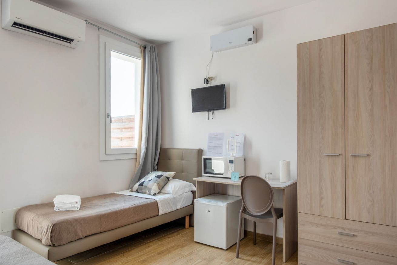 Ein Schlafzimmer mit einem Bett, einem Schreibtisch mit Computer, einem Sessel, einem Kleiderschrank und einer Klimaanlage.