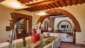 A dining area with a glass table, red vases, and a brick archway leading to a living room with a sofa and a TV. - Podere Pozzangone - Your Tuscany Home (Lucignano)