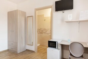 Habitación con armario, microondas, escritorio, silla, televisor y zona de ducha.
