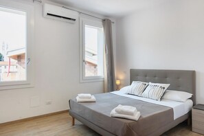 Un dormitorio con una cama, dos ventanas, un aire acondicionado, una mesita de noche y una lámpara.