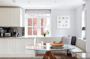 A modern kitchen with white cabinets, a glass-top dining table, and a view of a brick building through a large window.