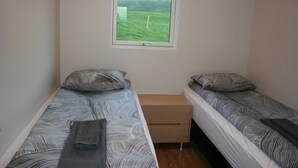 A small room with two single beds, each with patterned bedding and pillows. There is a small wooden nightstand between the beds. A window above the beds offers a view of a green landscape. - Eyvindartunga Farm Cottage (Bláskógabyggd)
