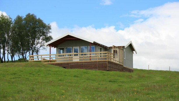 A single-story cabin with a wooden deck and a small porch, surrounded by a grassy field and trees. - Eyvindartunga Farm Cottage (Bláskógabyggd)