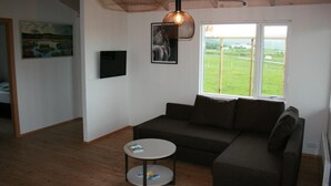 A modern living room with a sectional sofa, a round coffee table, and a large window offering a view of a green landscape. - Eyvindartunga Farm Cottage (Bláskógabyggd)