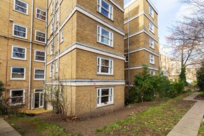 A multi-story brick building with multiple windows, a visible fire escape, and a pathway leading to the entrance. - Beautiful 2 Bedroom Garden flat (London)