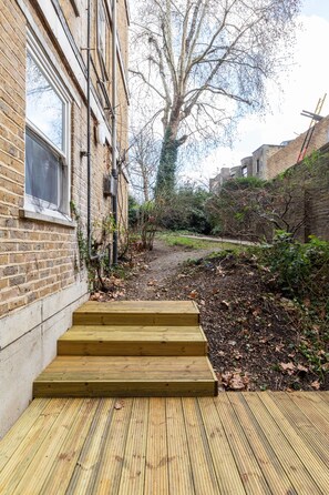A wooden staircase leading to a brick building with a window and a tree in the background. - Beautiful 2 Bedroom Garden flat (London)