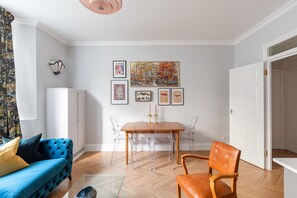 Een moderne woonkamer met een blauwe bank, een houten eettafel met transparante stoelen, en een witte kast.