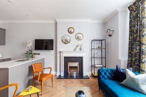 A modern living room with a fireplace, a blue sofa, and a dining area with a small table and chairs. - Beautiful 2 Bedroom Garden flat (London)