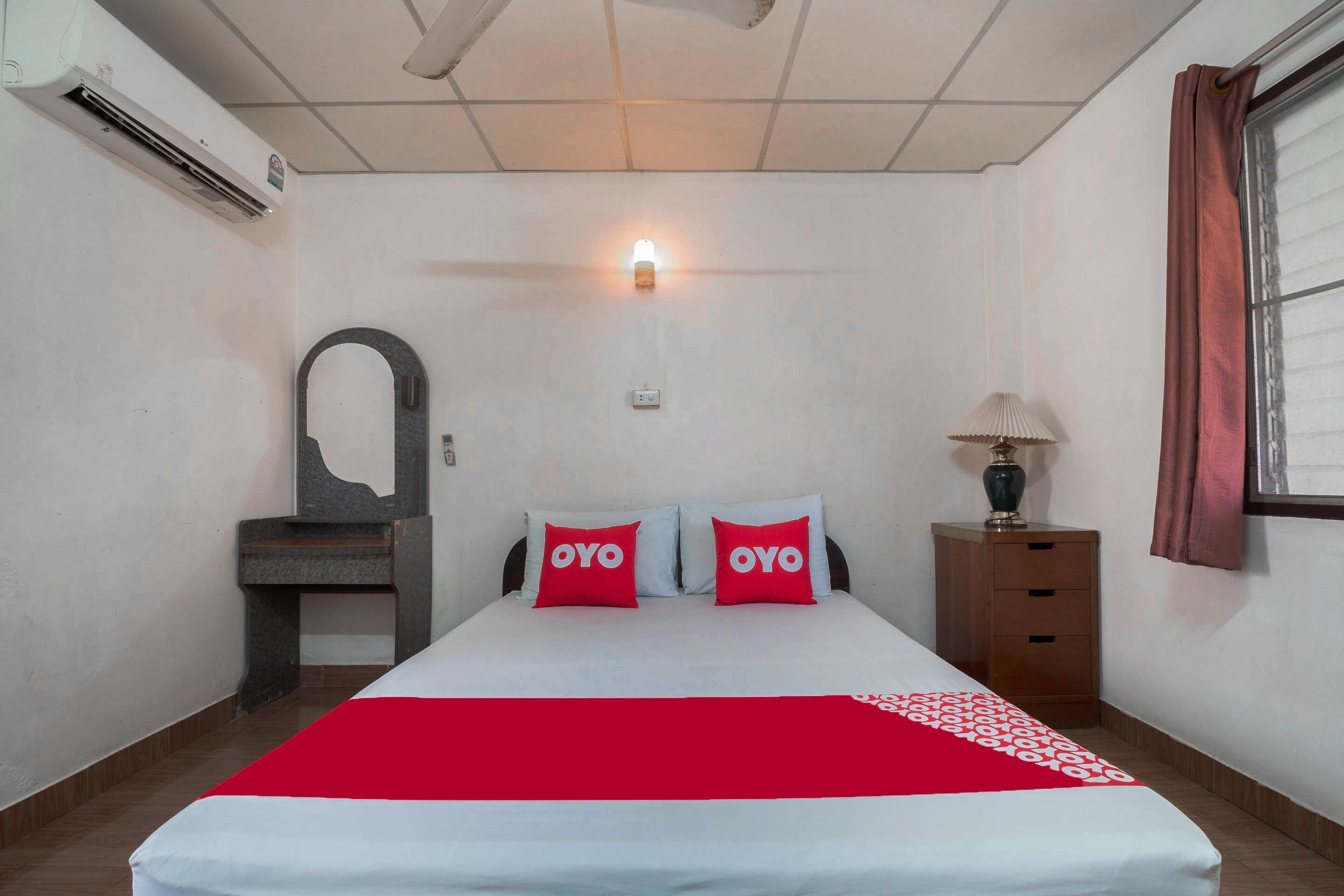 Une chambre d’hôtel comprenant un lit, deux oreillers portant la marque « OYO », une lampe de chevet, une table de chevet et un appareil de climatisation.