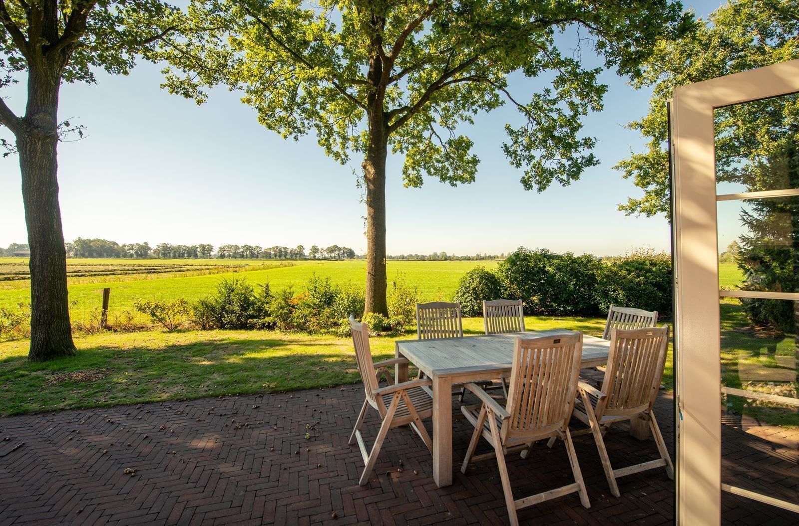 An outdoor patio with a wooden table and chairs, surrounded by trees and a view of a green field.