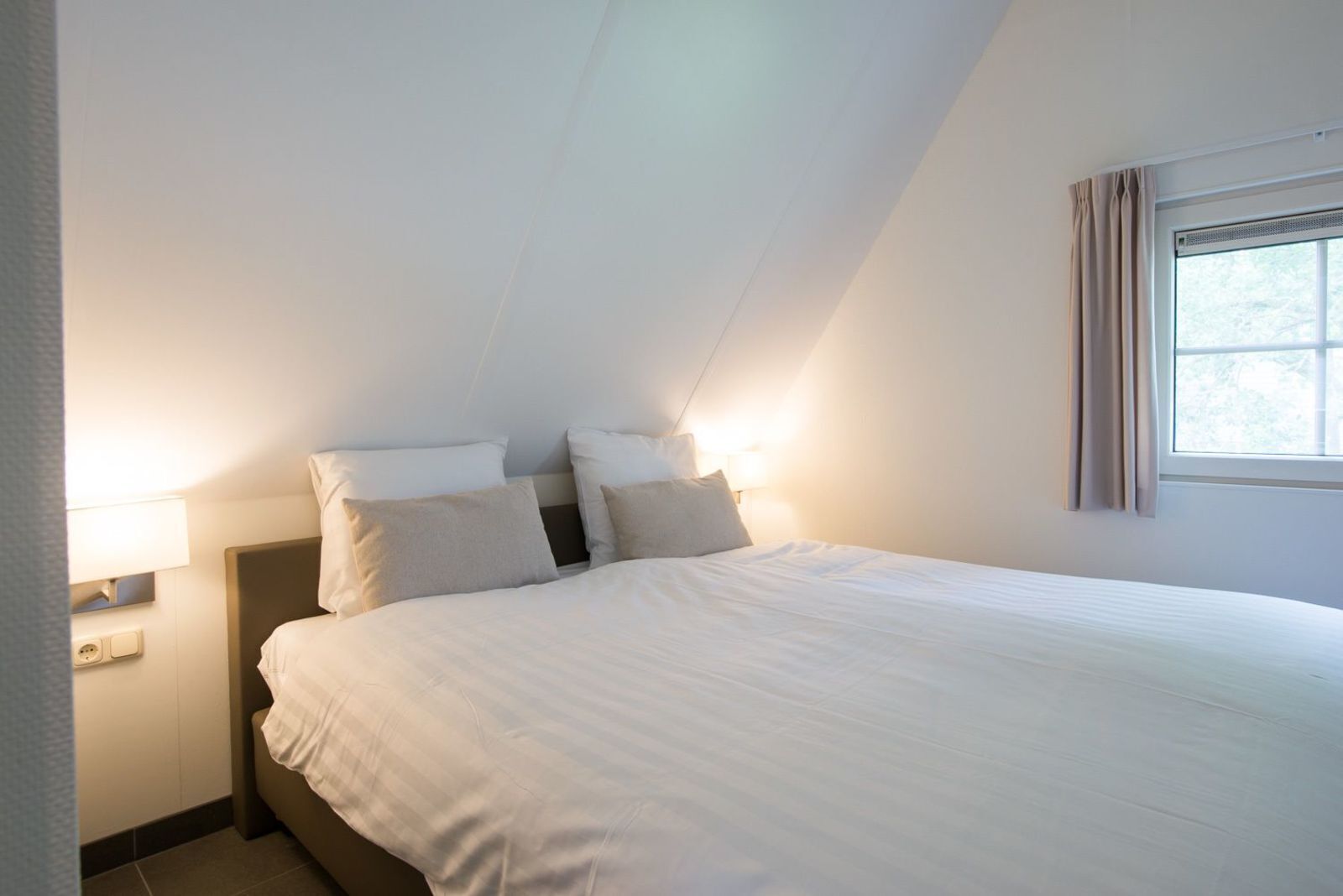 Quarto com cama, duas luminárias de cabeceira, janela com cortinas e uma tomada.