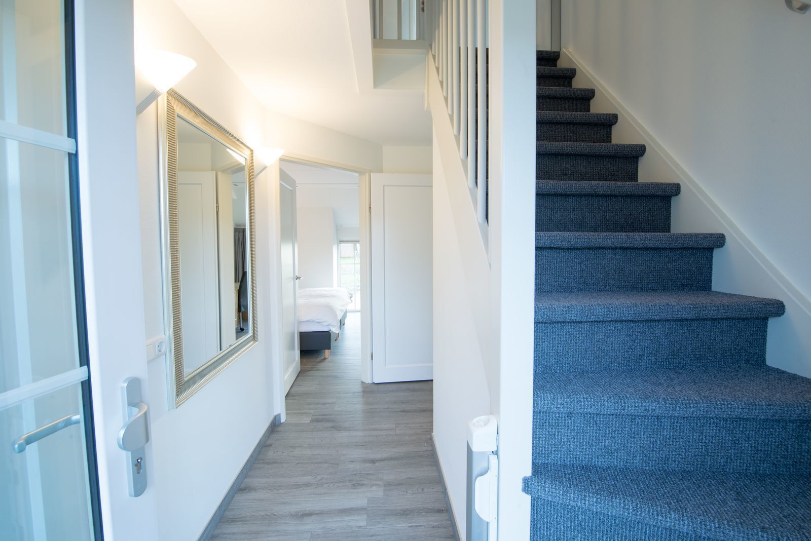 A hallway with a staircase leading to a bedroom. The hallway has a mirror, a door, and a light fixture. The staircase has carpeted steps.