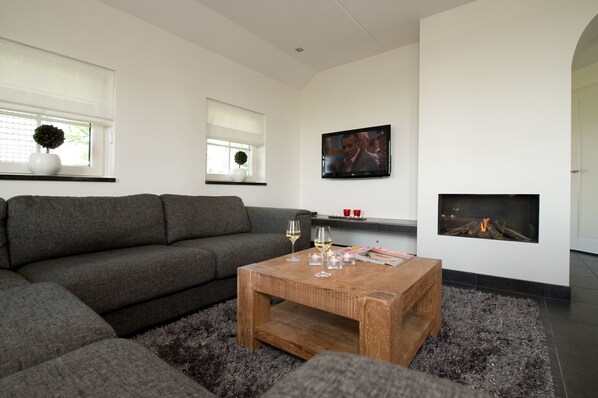Una sala moderna con un sofá seccional gris, una mesa de centro de madera con vasos y velas, un televisor de pantalla plana y una chimenea empotrada.