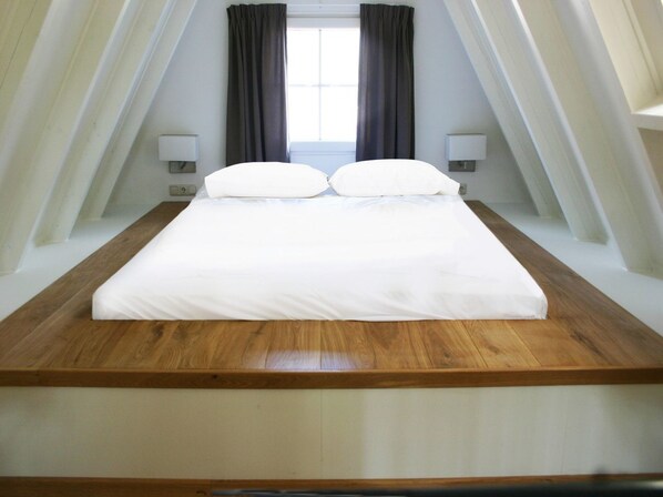 Un dormitorio con cama plataforma de madera, ropa de cama blanca y ventana con cortinas.