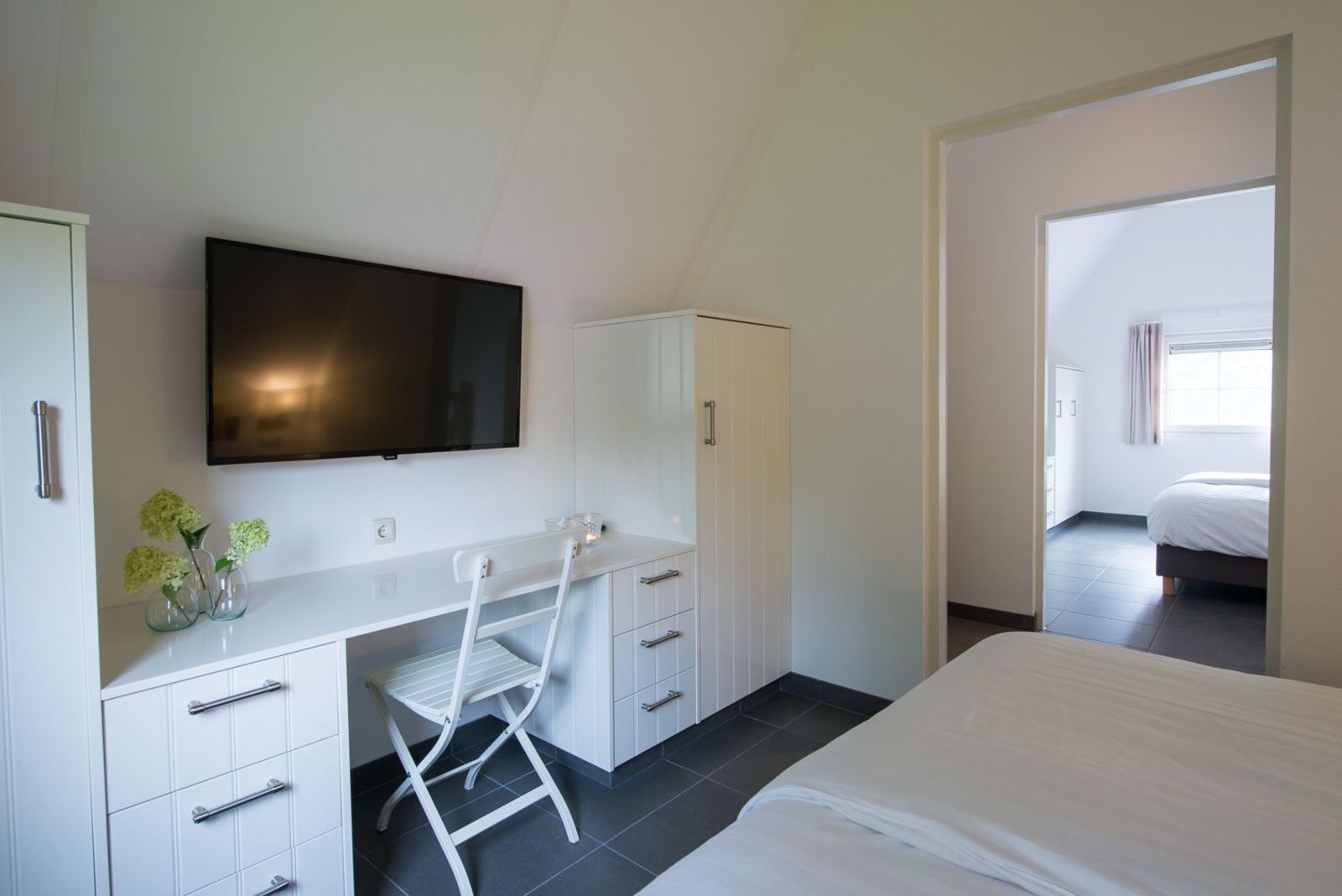 Ein modernes Schlafzimmer mit einem Bett, einem Schreibtisch mit Schubladen, einem Stuhl, einem an der Wand montierten Fernseher und einem Fenster.