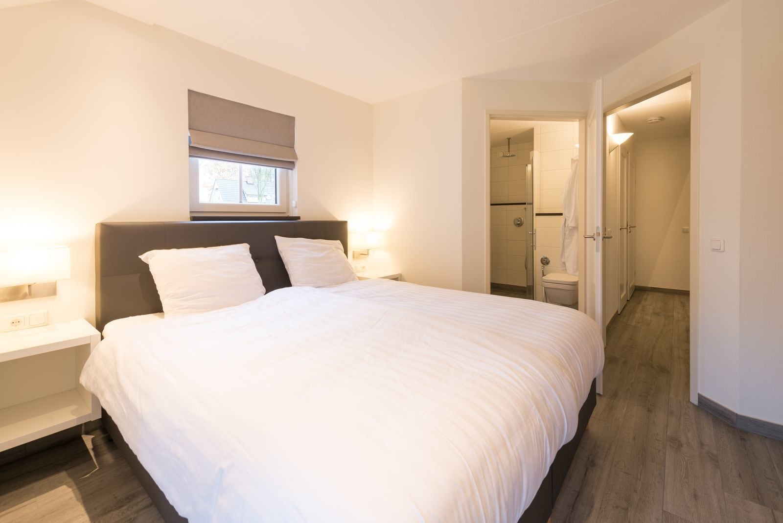 A modern bedroom with a large bed, bedside tables, a window with a blind, and a bathroom visible through an open door.