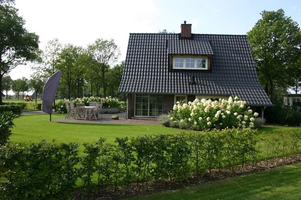 A house with a dark tiled roof, a brick exterior, and a front yard with a lawn, a patio with chairs, and a large white flower bush. - Hof van Salland (Hellendoorn)