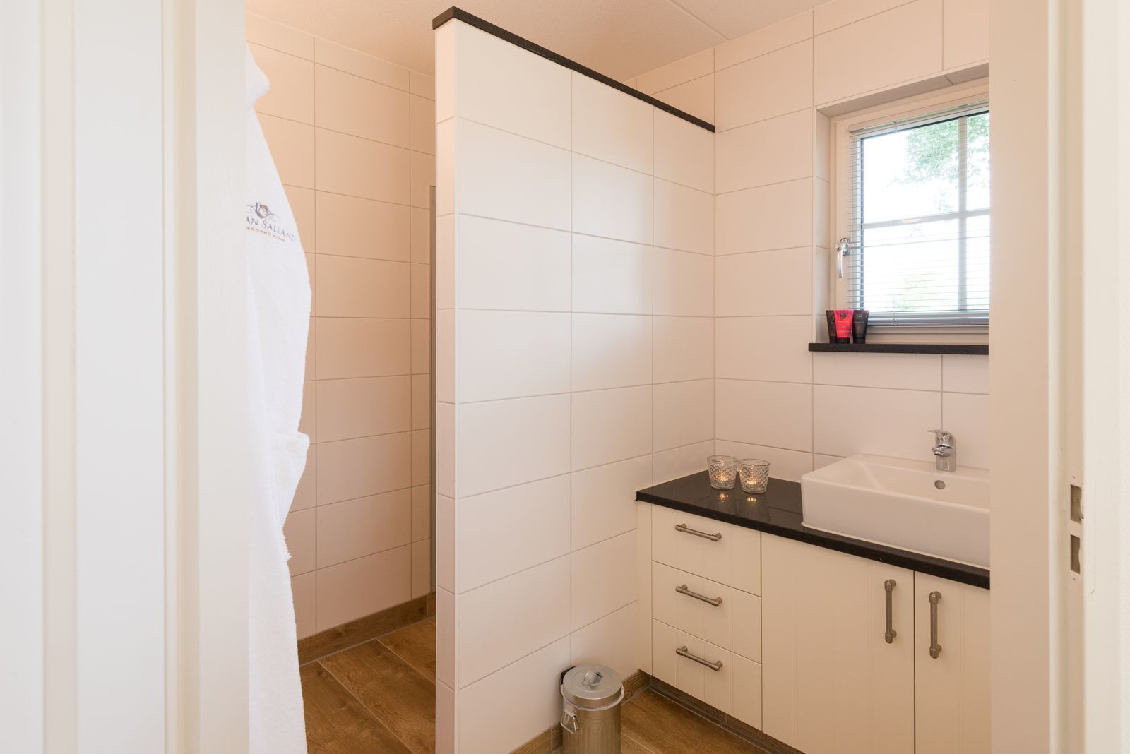 A bathroom with white tiles, a black countertop, a window, and a small trash bin.