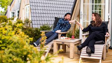 Een koppel zittend aan een houten tafel met een glas wijn, onder een grote parasol, in een gezellige buitenomgeving met houten meubels en een pannendak.