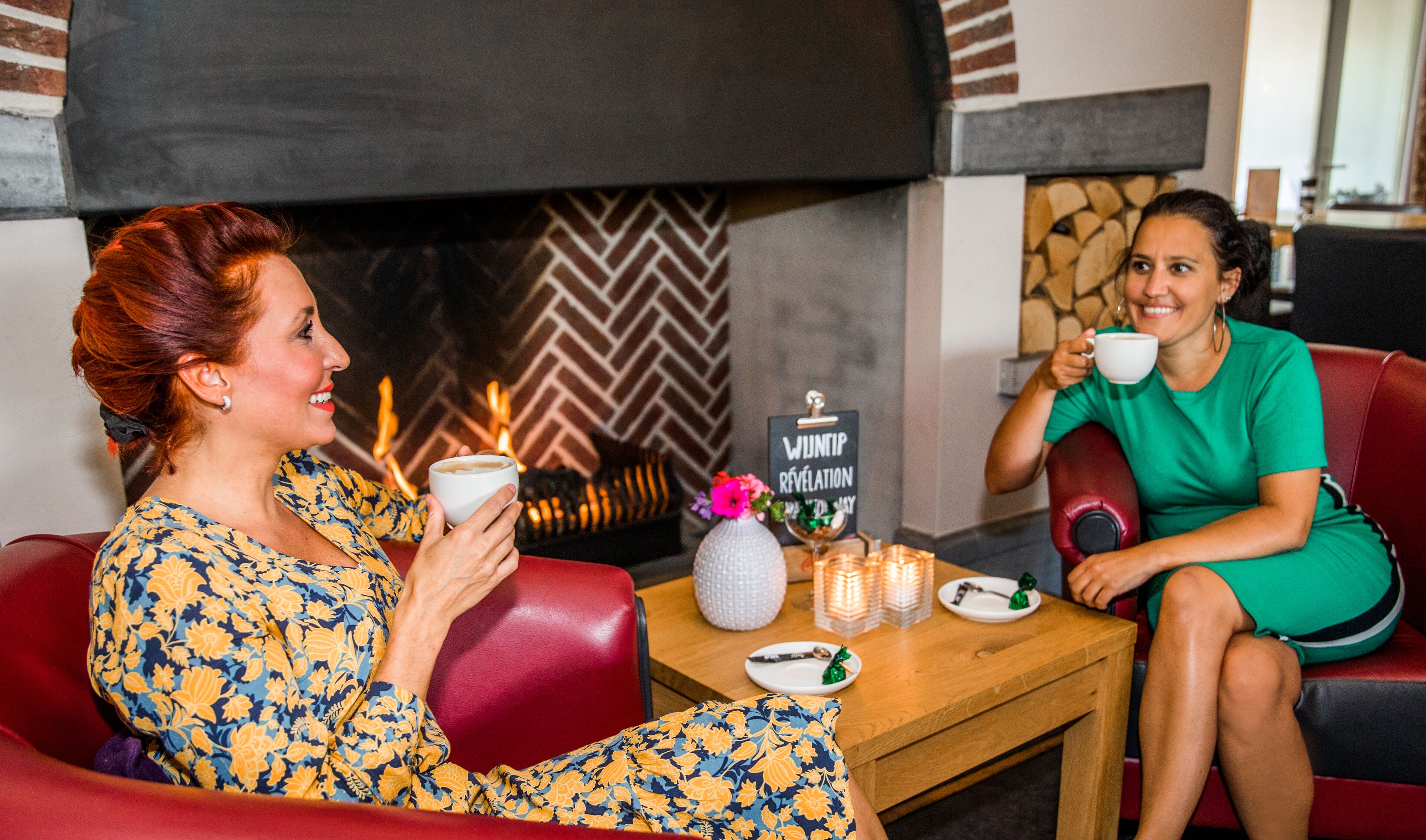 Two people sitting on red leather sofas in a cozy room with a fireplace, a small table with a vase and candles, and a sign reading "WUNTIP RÉVÉLATION".