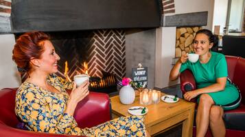 Two people sitting on red leather sofas in a cozy room with a fireplace, a small table with a vase and candles, and a sign reading "WUNTIP RÉVÉLATION".