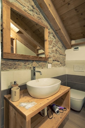 A bathroom with a stone wall, wooden beams, a large white sink, and a toilet. - Casa Ekharle - Guesthouse (Rotzo)