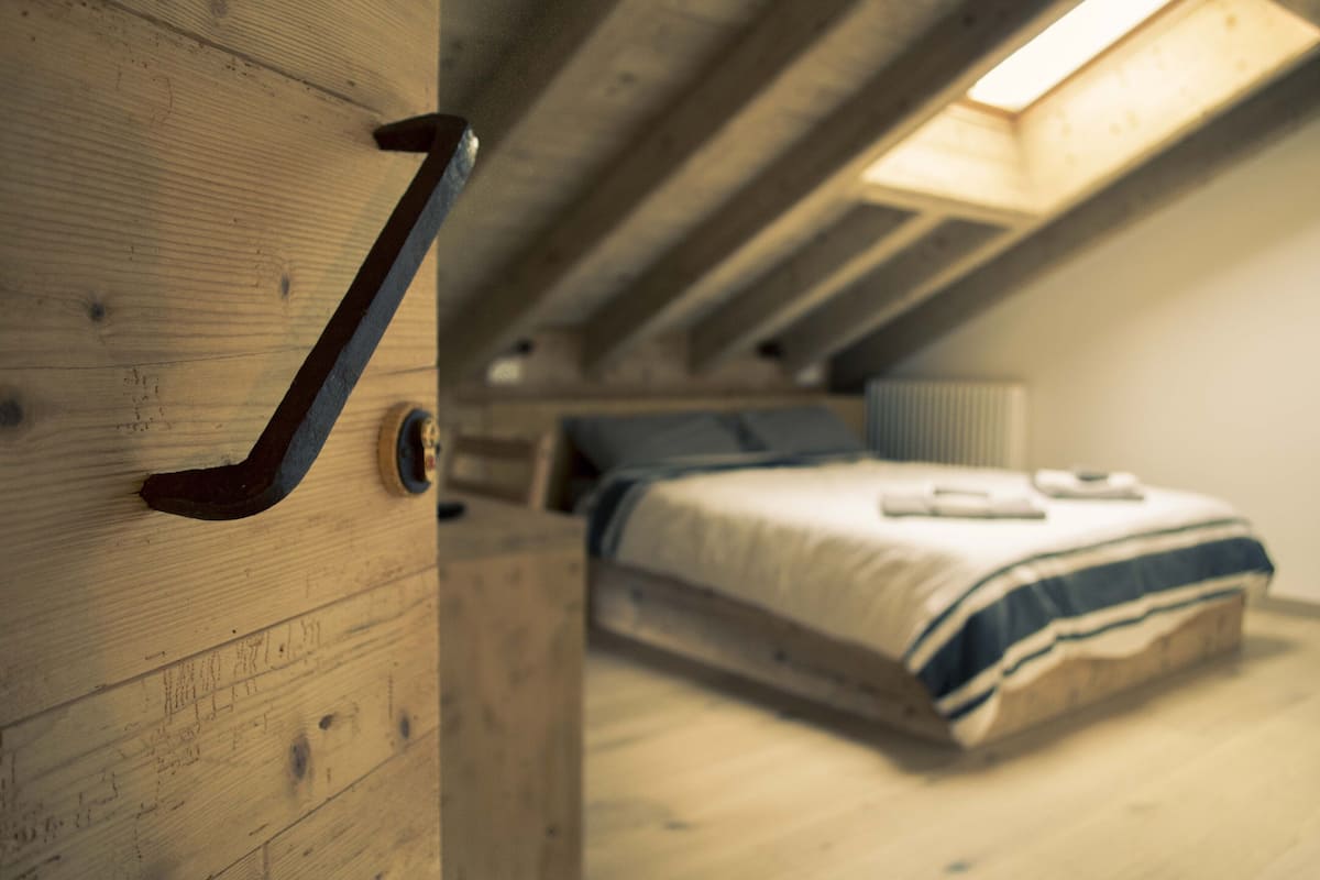 Quarto com teto inclinado, cama com manta listrada e porta de madeira com puxador.