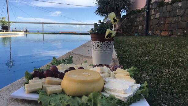 A cheese platter with various cheeses and grapes on a white plate, placed on a concrete surface near a pool with a view of a bridge and palm trees in the background. - Old kitchen Cus cus factory Villa with pool and sea view (Funchal)