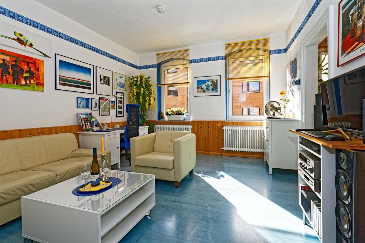 A living room with a sofa, armchair, coffee table, and a small kitchen area with a washing machine.