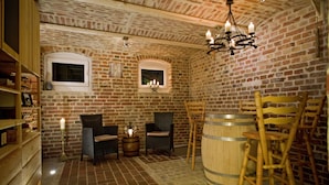 Una habitación rústica con paredes de ladrillo, muebles de madera y una araña. Se observan dos ventanas, un barril de madera y una estantería con objetos.