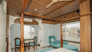 A hotel room with a bed, a table, two chairs, a window with a view of a river and buildings, and a ceiling fan. - Jheel Hotel and Restaurent (Udaipur)