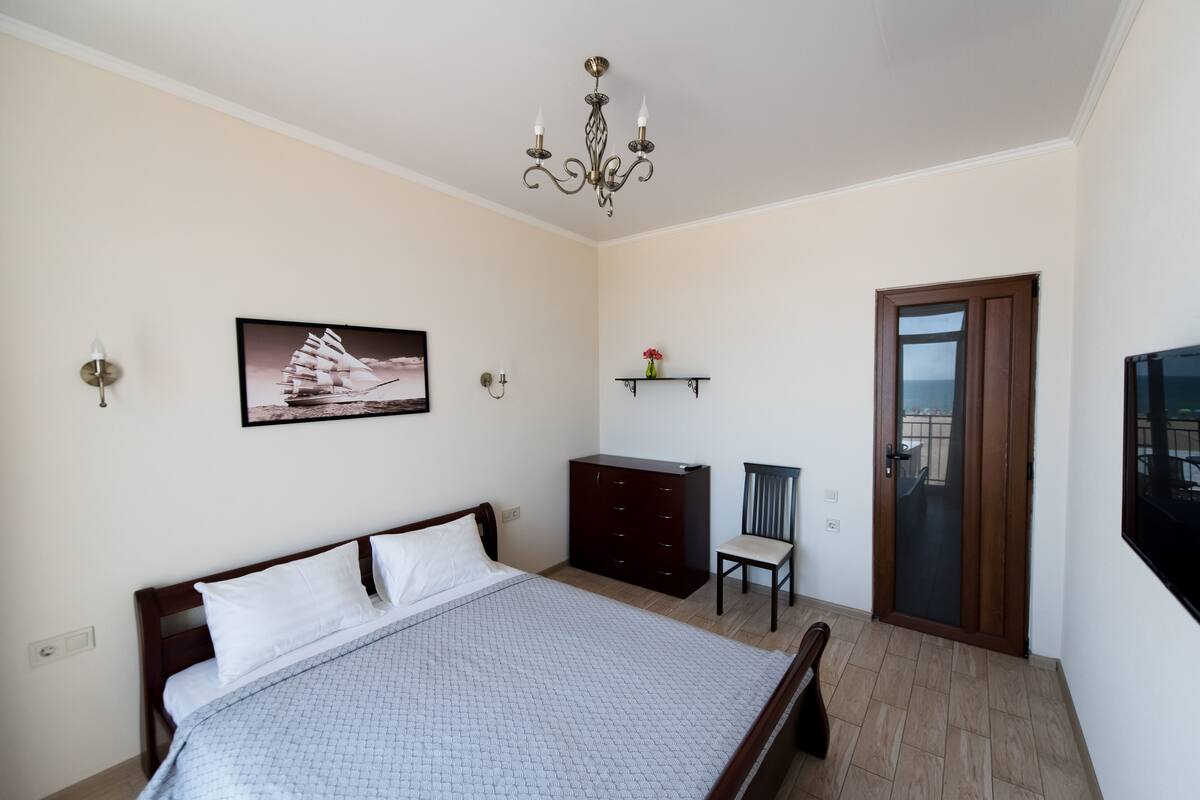 Een slaapkamer met een bed, een dressoir, een stoel, een deur, een kroonluchter en een aan de muur bevestigd schilderij.