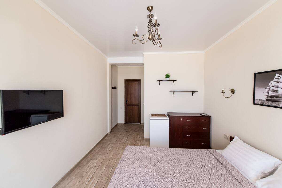 Een slaapkamer met een bed, een dressoir, een aan de muur bevestigde televisie, een kroonluchter en een deur die naar een andere kamer leidt.