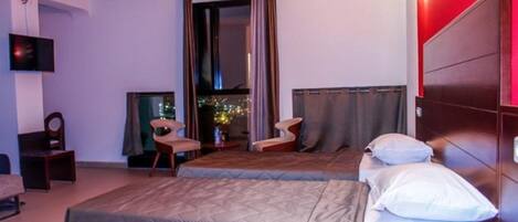A hotel room with a bed, two chairs, a TV, and a view of a cityscape through a window.