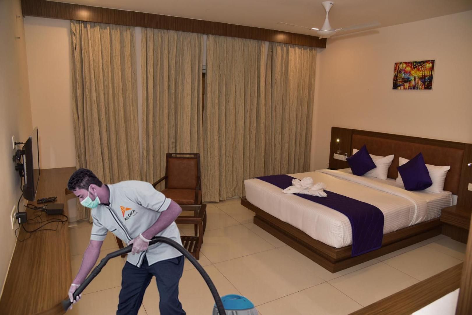 A hotel room with a bed, a television, a chair, and a person cleaning.